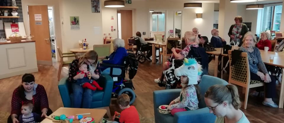 This picture shows babies, toddlers, parents, carers and older people in a care home sitting and playing together at Leeds NCT's intergenerational playgroup, held in a care home