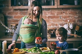 A mother preparing food with baby in a sling and a toddler