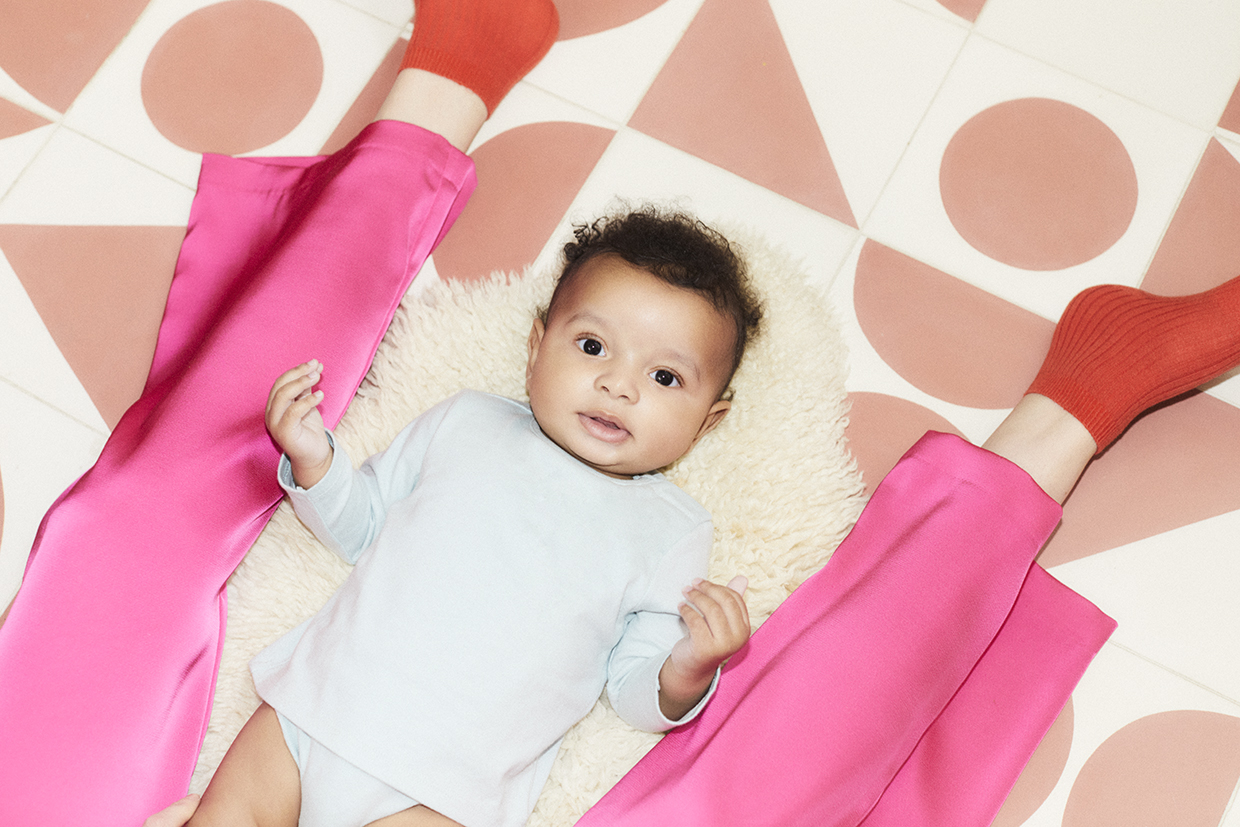 Baby lies on a neutral rug, between an adults legs looking up softly at the camera. The adult wears bright pink trousers and red socks as they sit on the floor with the baby on a bright tiled floor