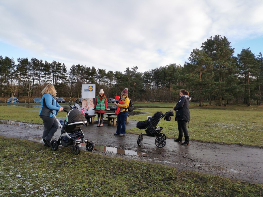 A group of parents standing with their pushchairs in a park next to an NCT banner