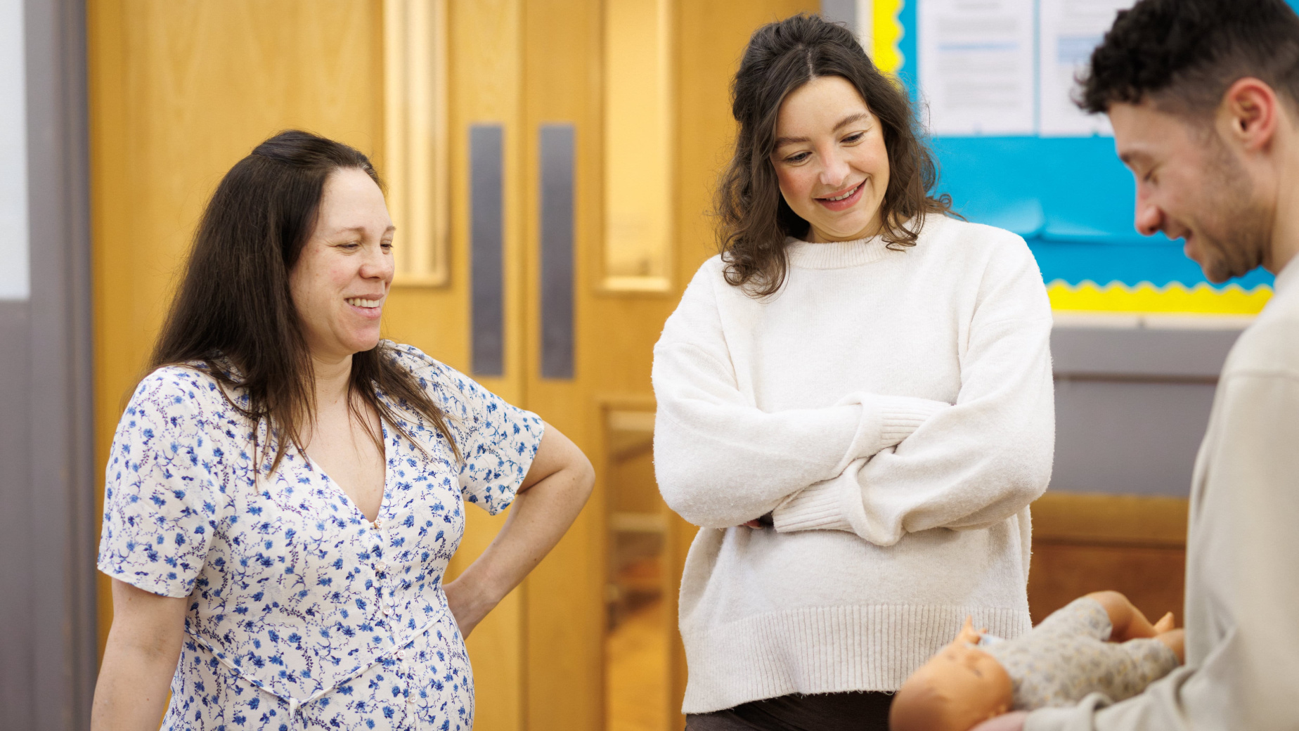 Two pregnant woman smiling and looking at dad holding a doll at an antenatal class