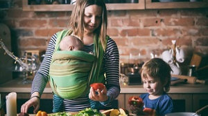 A mother preparing food with baby in a sling and a toddler