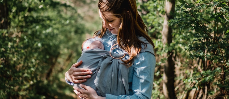 A woman on a sunny forest path, carrying a baby in a ring sling