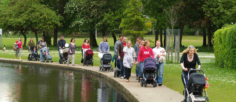 Image of parents pushing pushchairs with a pond to their right and grass to their left. 