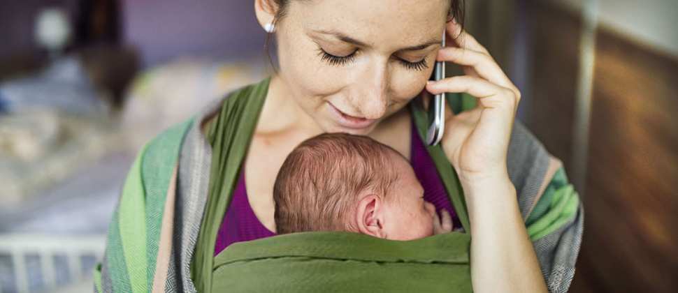 Head and shoulder of woman on phone her face turned down towards a newborn baby sleeping in a sling